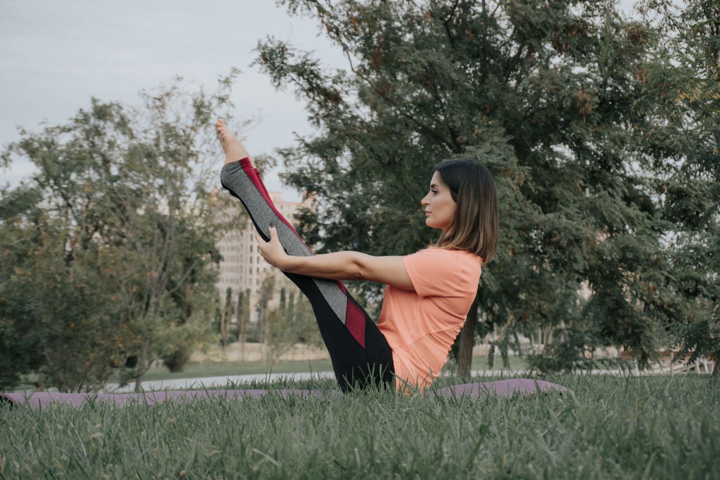 A woman practicing yoga outdoors in a park, performing a seated leg stretch on a mat.