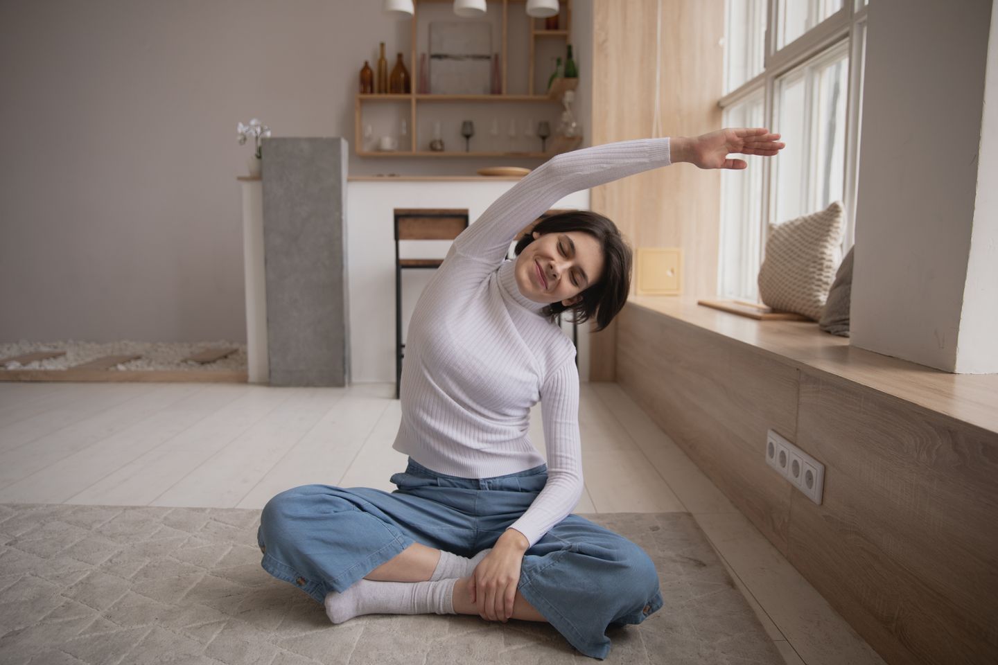 Young female in casual wear sitting with closed eyes and crossed legs on carpet while doing side inclination in apartment